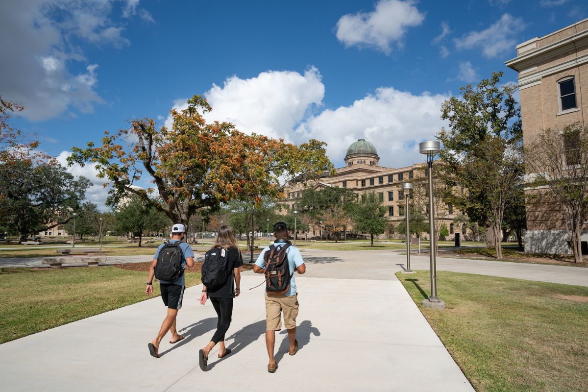 Three students with backpacks walk toward the academic building on a sunny day with blue sky and some clouds